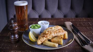 Un plato de pescado con papas fritas, una cerveza y salsas sobre una mesa de madera.