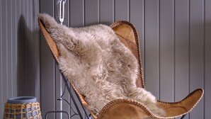 A chair with a fur-like cushion, a round table with a woven basket, and a floor lamp in a room with vertical paneling.