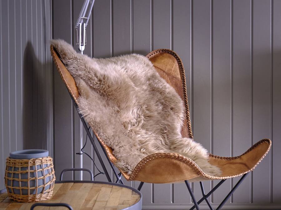 A chair with a fur-like cushion, a round table with a woven basket, and a floor lamp in a room with vertical paneling.