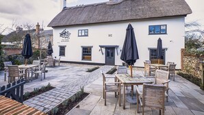 A white building with a thatched roof, numbered 1612, featuring outdoor seating with wooden tables and chairs, and umbrellas.