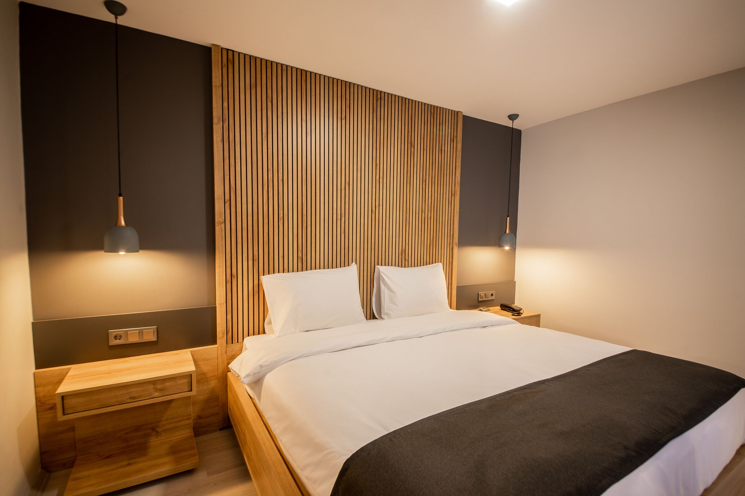 A modern bedroom with a wooden headboard, a large bed with white bedding, two bedside tables, and pendant lighting.