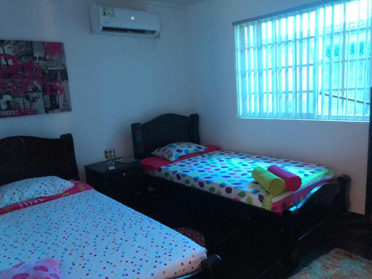 Quarto com duas camas, uma mesinha de cabeceira, ar-condicionado fixado na parede e uma janela com persianas.