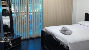 Habitación de hotel con cama, espejo, televisión y vista a una piscina a través de persianas verticales.
