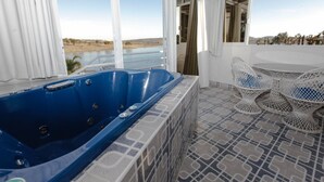 Una habitación con un jacuzzi azul grande, piso estampado y vistas a un cuerpo de agua y colinas.