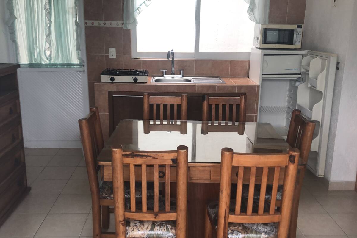 Een kleine keuken met een eettafel en stoelen, een kookplaat, een spoelbak en een magnetron.