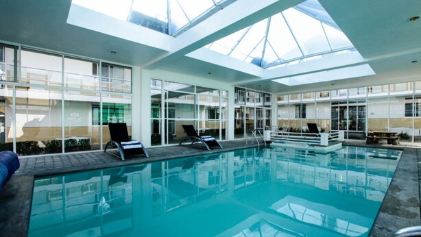 Una piscina interior con techos de vidrio transparente, sillones de descanso y vistas del edificio exterior.