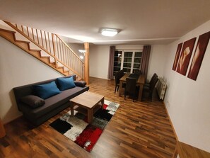 Un salon avec un escalier en bois, un canapé avec des coussins, une table basse, une table à manger avec des chaises et un radiateur.