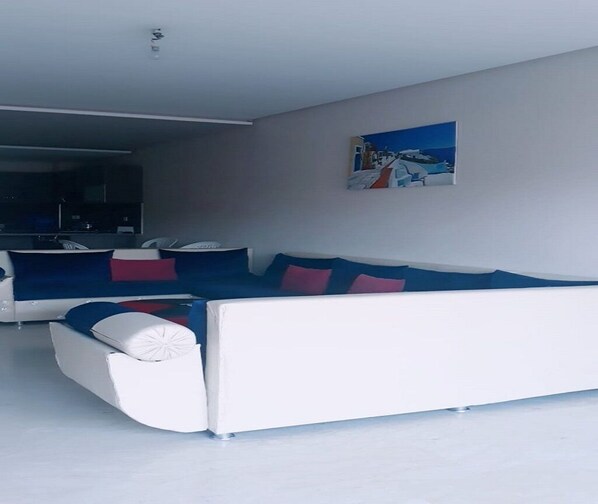 A modern living room with a white sofa and blue cushions, a painting on the wall, and a kitchen area in the background.