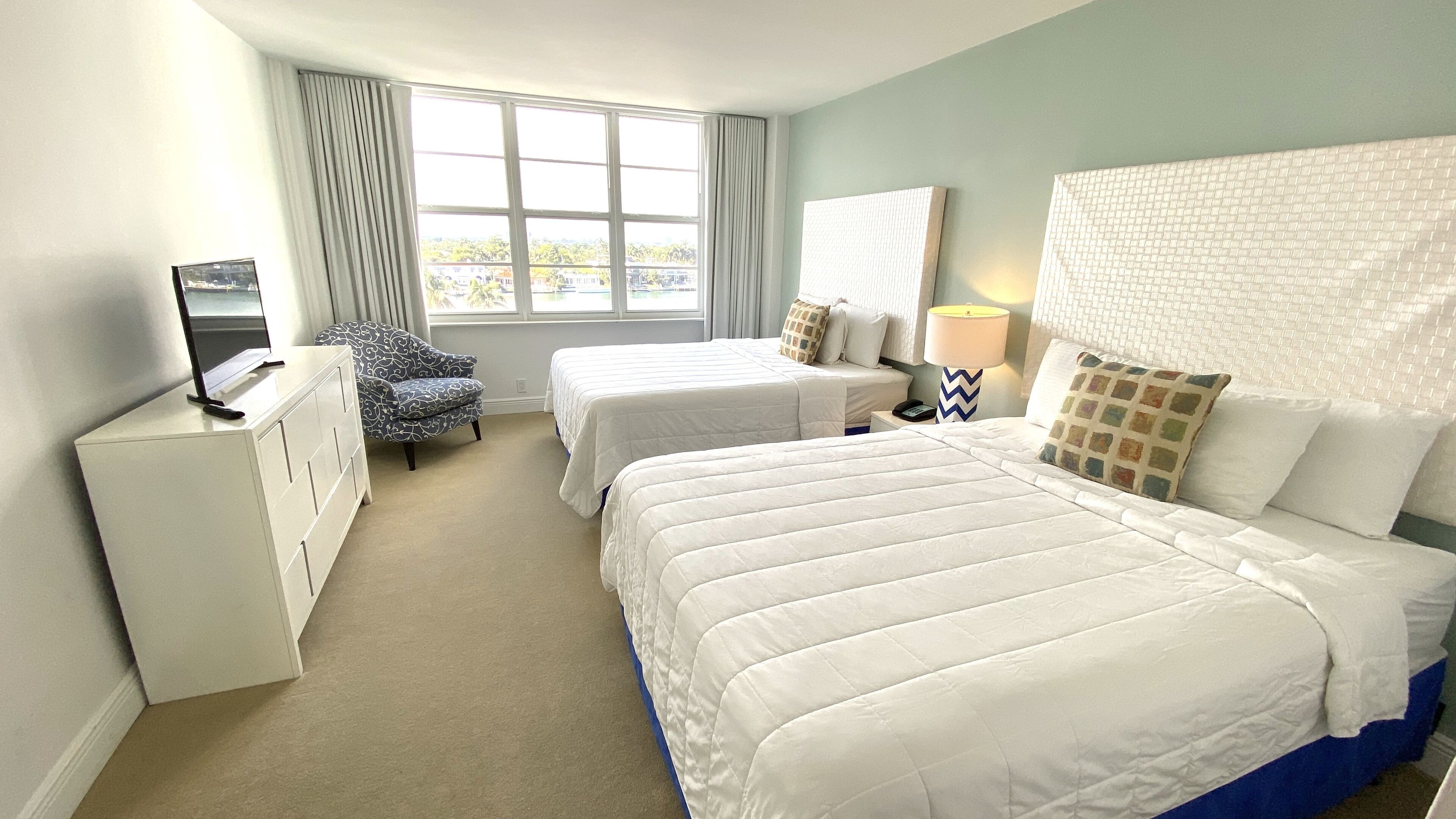 Ein Hotelzimmer mit zwei Betten, einem Sessel, einem Schreibtisch, einer Lampe und Blick ins Freie.