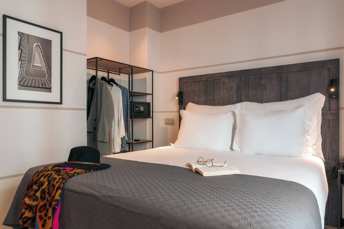 A neatly made bed with white pillows and a gray blanket, a black hat, and a pair of glasses on the bed.