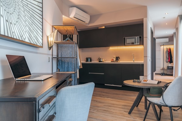 A modern kitchen with dark cabinets, a microwave, and a coffee machine. There is a laptop on a desk, a chair, and a table with a hat and sunglasses.
