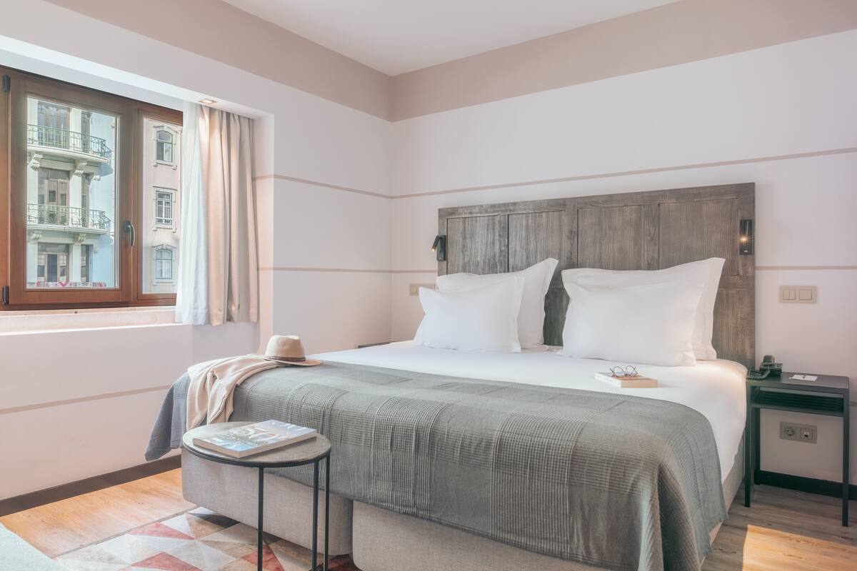 A bedroom with a large bed, a bedside table, a window with curtains, and a view of buildings outside.
