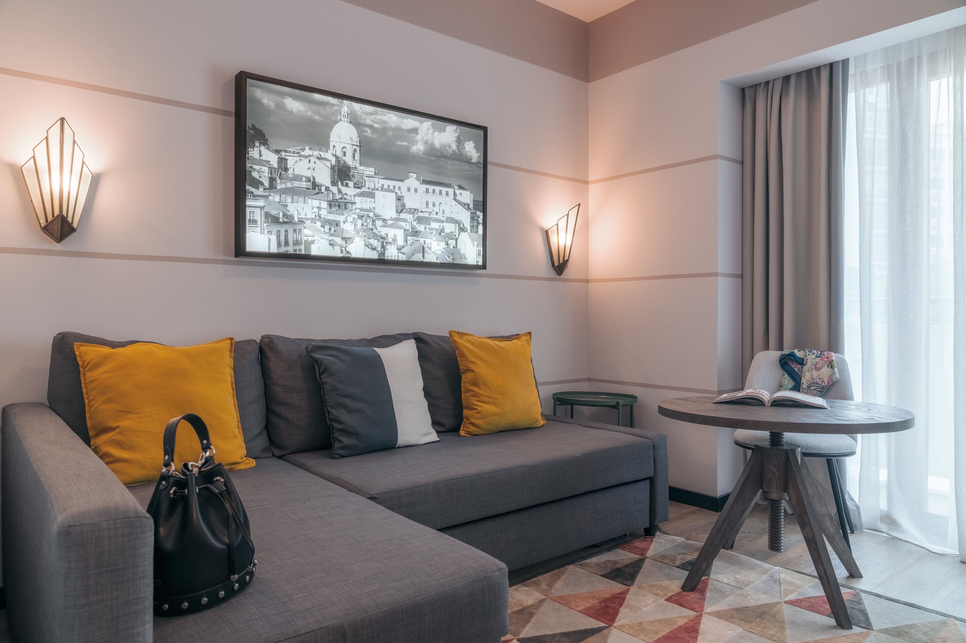 Un hotel moderno con un sofá gris, cojines amarillos y grises, un bolso negro, una mesa redonda de madera con un libro, y una imagen enmarcada de un paisaje urbano en la pared.
