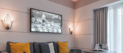Un hotel moderno con un sofá gris, cojines amarillos y grises, un bolso negro, una mesa redonda de madera con un libro, y una imagen enmarcada de un paisaje urbano en la pared.