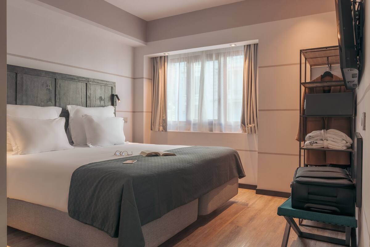 A neatly made bed with a grey headboard, a bedside table with a book and glasses, a window with sheer curtains, and a luggage rack against the wall.