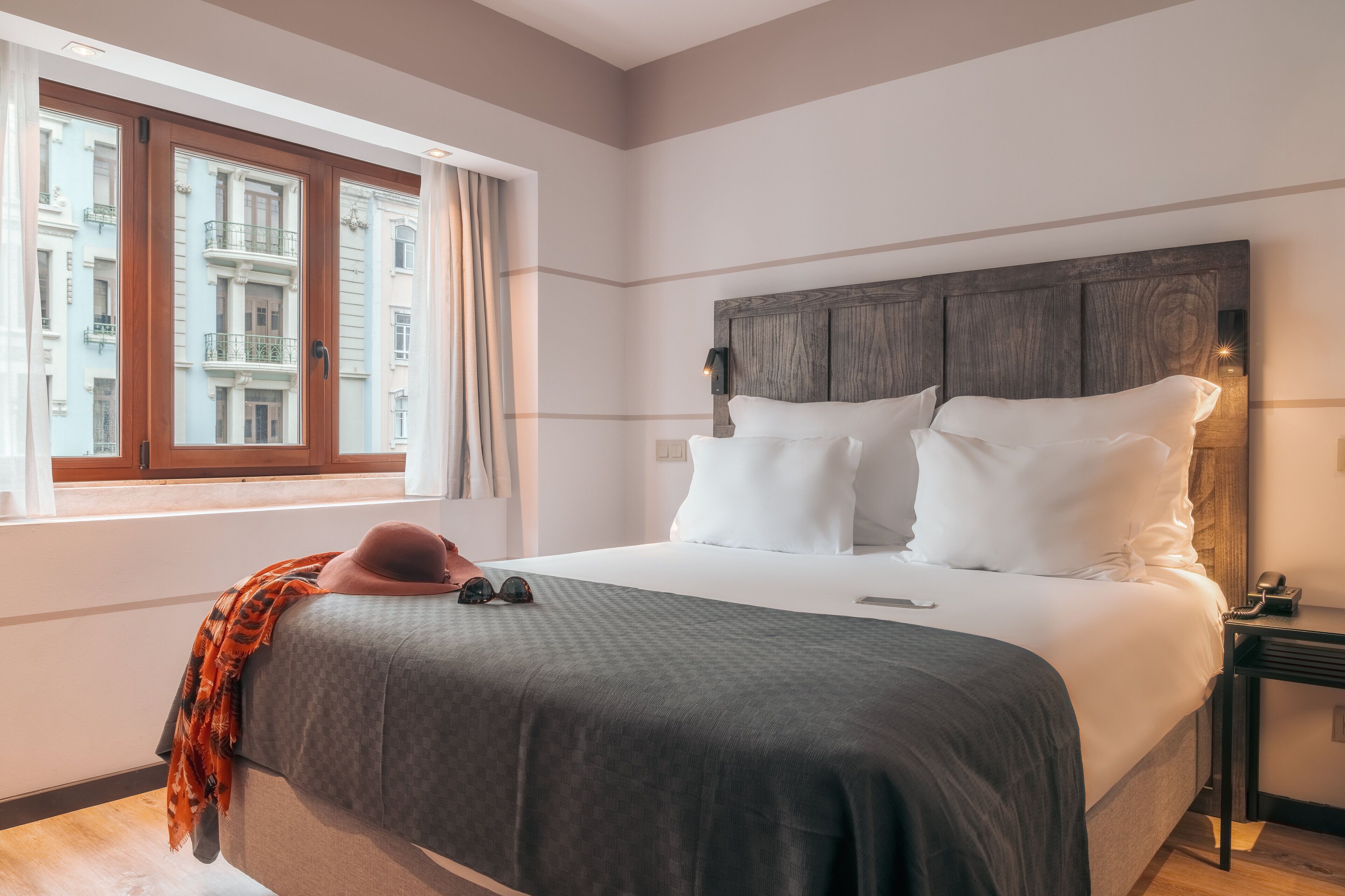 A bedroom with a large bed, a nightstand, a window with curtains, and a view of buildings outside.