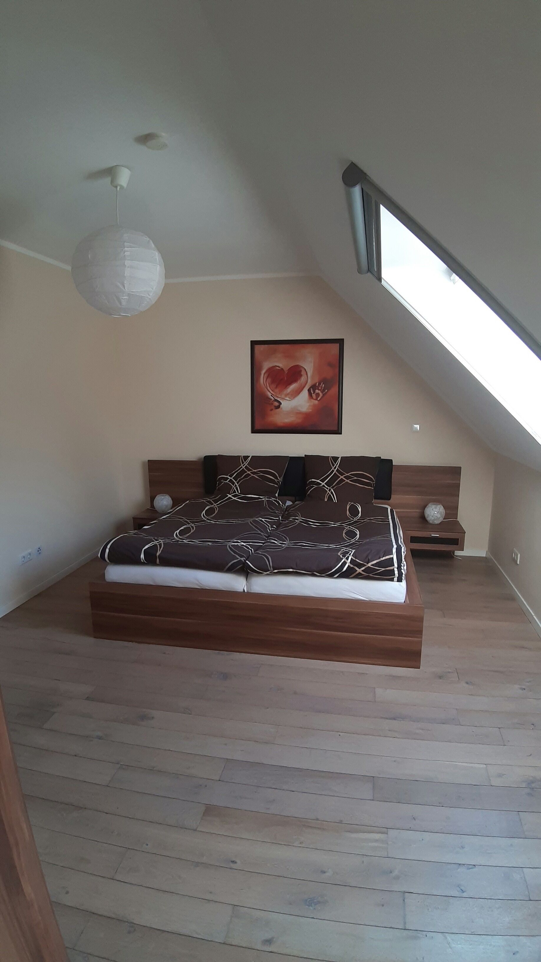 Ein Schlafzimmer mit schrägen Wänden, einem Bett mit gemustertem Überwurf, einem gerahmten Bild an der Wand und einem Oberlicht.