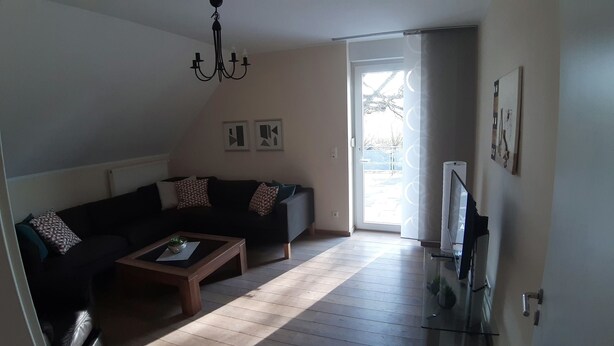 A living room with a dark-colored sofa, a wooden coffee table, and a chandelier. There is a door leading to a balcony with a view of trees. - Apartment right on the Dutch border (Oeding)