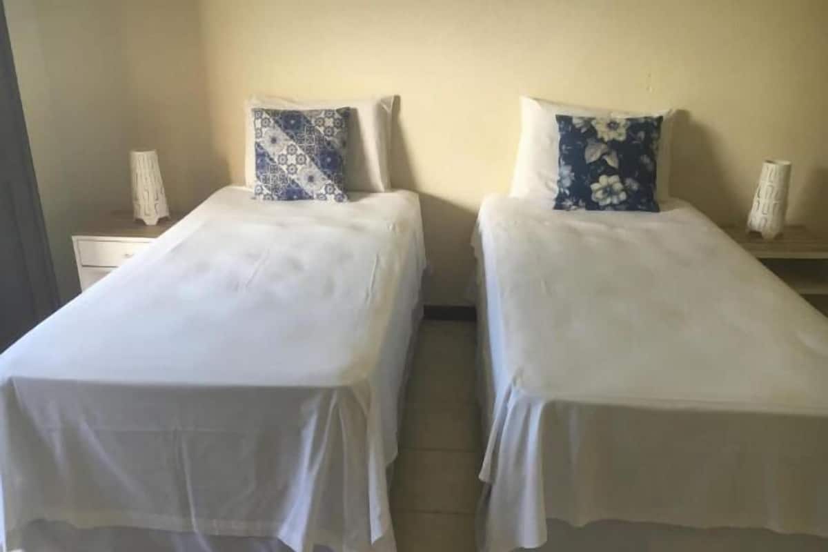 Dos camas individuales con sábanas blancas y almohadas estampadas, una mesita de noche con lámpara, y una pared lisa.