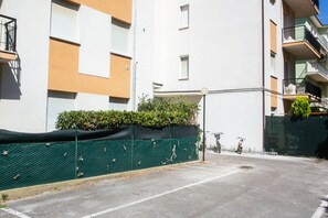 Un espace de stationnement délimité par une clôture verte, un bâtiment doté de balcons et un ciel dégagé.