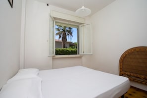 A bedroom with a single bed, a window with a view of a palm tree and building, and a wicker chair.