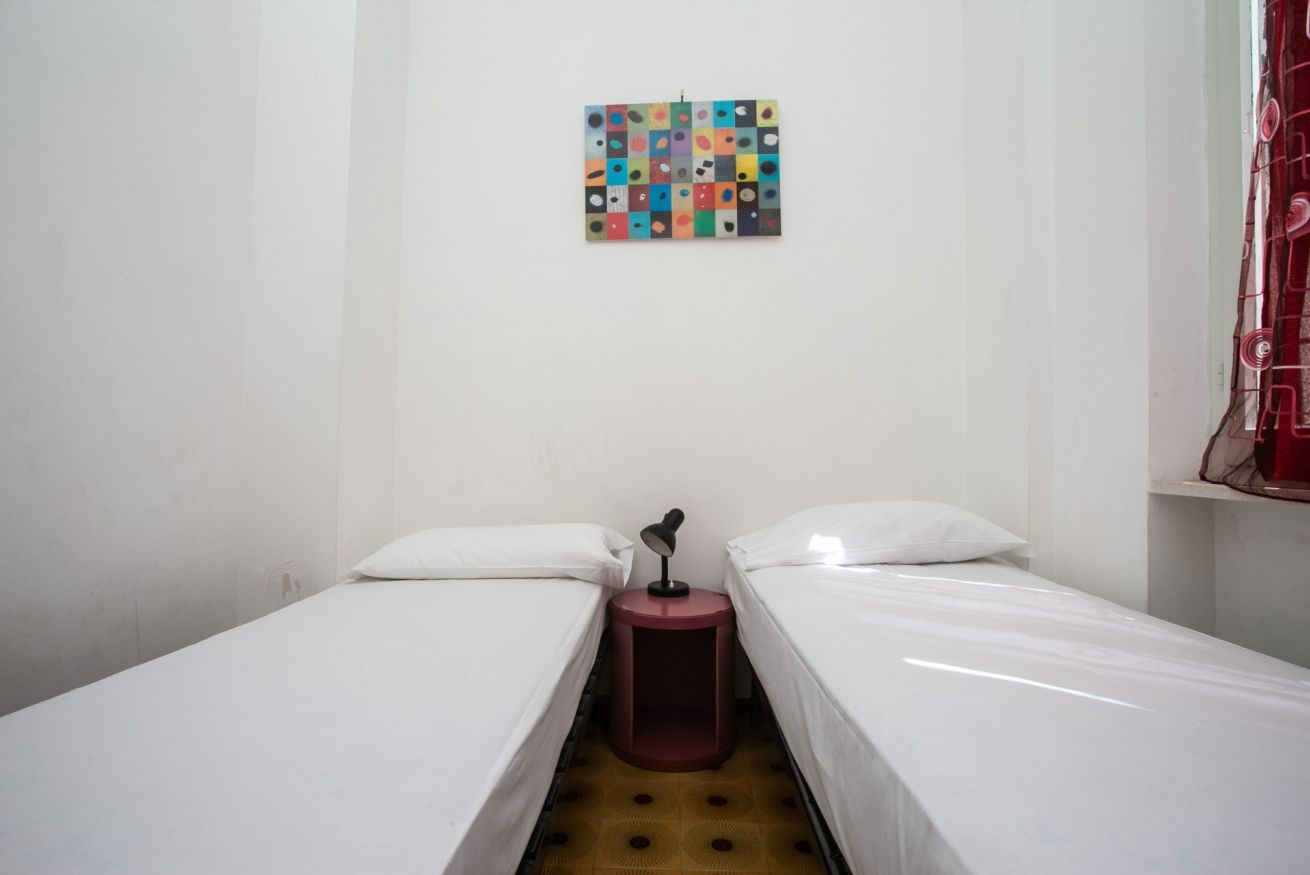 A small, clean room with two single beds, a small table with a lamp, and a colorful abstract painting on the wall.