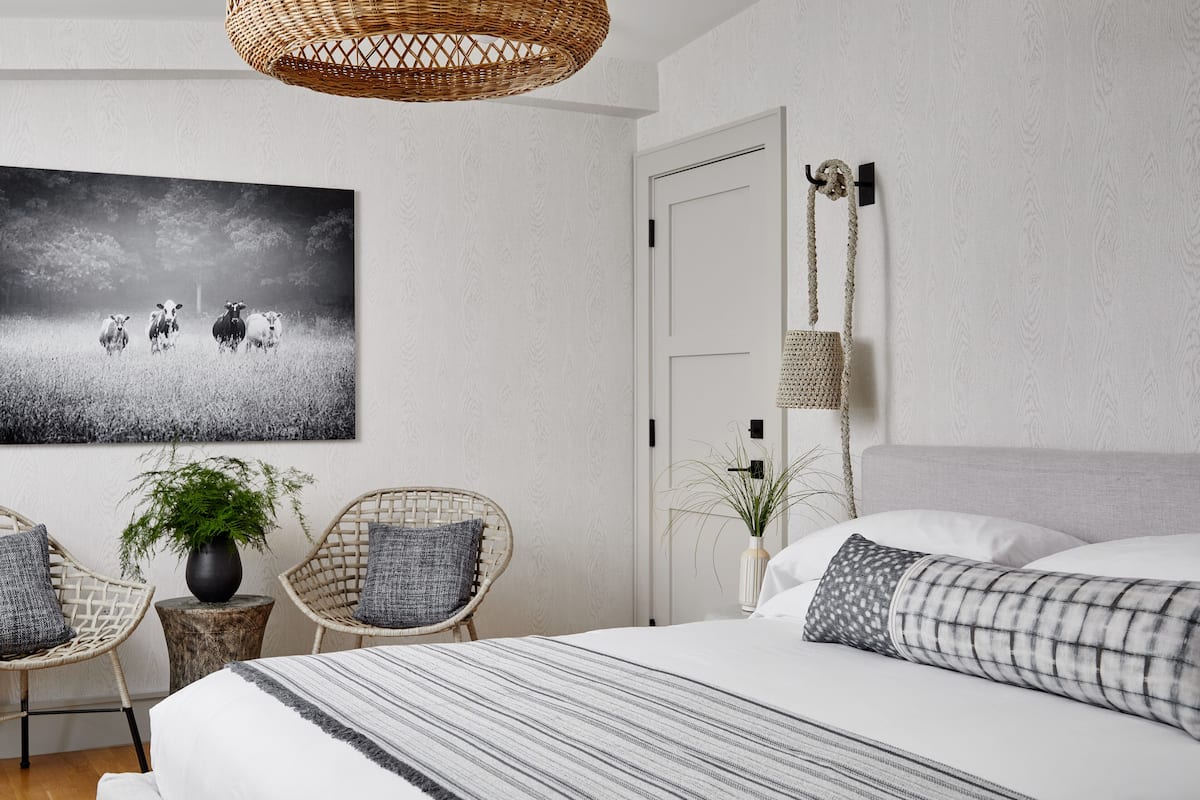 A bedroom with a large bed, two wicker chairs, a potted plant, a wall-mounted lamp, and a framed picture.
