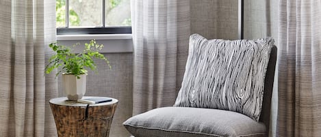 Ein Zimmer mit Fenster, Vorhängen, einem Nachttisch mit Pflanze, einem Sessel mit gemustertem Kissen und einer Pendelleuchte.