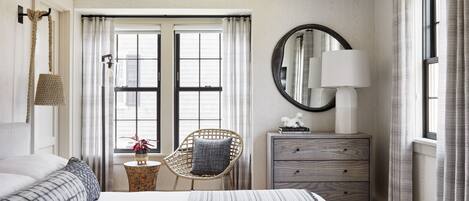 A hotel room with a large bed, a wicker chair, a dresser, and a window with curtains.