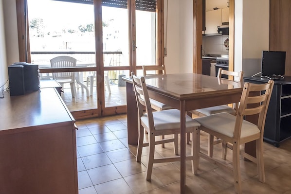 A dining area with a wooden table and chairs, a kitchen in the background, and a view through a glass door.
