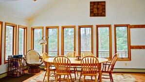 A dining area with a round table and six wooden chairs, large windows with a view of trees, and a lamp on a side table. - Lynch Mountain Retreat (Cooperstown)