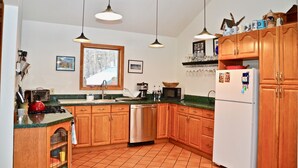 A kitchen with wooden cabinets, a stainless steel dishwasher, a microwave, and a refrigerator. There are hanging lights and a window with a view of trees. - Lynch Mountain Retreat (Cooperstown)
