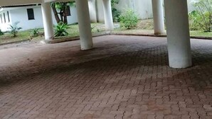 An open covered area with a brick floor and white pillars, surrounded by greenery and a building in the background. - 3 Bedroom Apartment In Nyali (Mombasa)