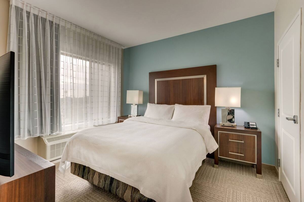 Une chambre d’hôtel avec un grand lit, deux tables de chevet avec des lampes, une télévision et une fenêtre avec des rideaux légers.