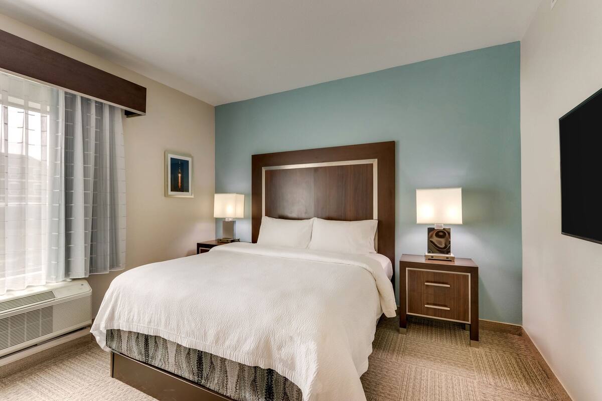 Une chambre d’hôtel avec un grand lit, deux tables de chevet avec des lampes, une fenêtre avec des rideaux et un téléviseur fixé au mur.
