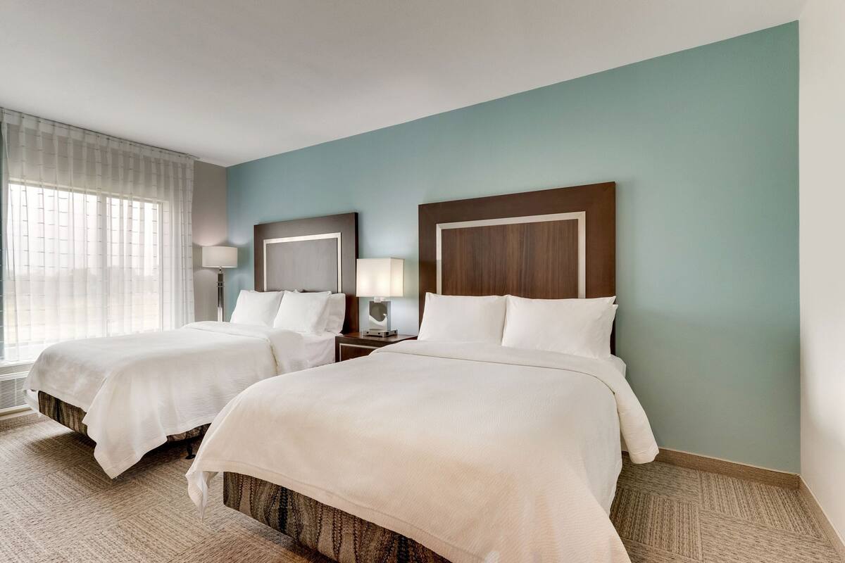 Une chambre d’hôtel avec deux lits, une grande fenêtre avec des rideaux légers, une table de chevet avec une lampe et un mur bleu clair.