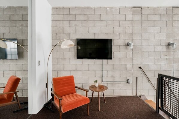 Une chambre moderne avec un mur en briques, un téléviseur à écran plat, une table ronde agrémentée d’une plante et deux chaises orange.