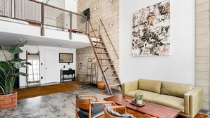 Un vaste espace de vie doté d’un haut plafond, d’un escalier en bois, d’un canapé beige, d’une table basse en bois et d’un grand tableau abstrait accroché au mur.