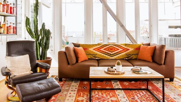 En moderne stue med en brun sofa, en læderstol, et sofabord og et stort vindue med udsigt til bygninger.