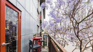 Ein Balkon mit einem roten Tisch und Stühlen, einer Topfpflanze und Blick auf blühende, violette Bäume.
