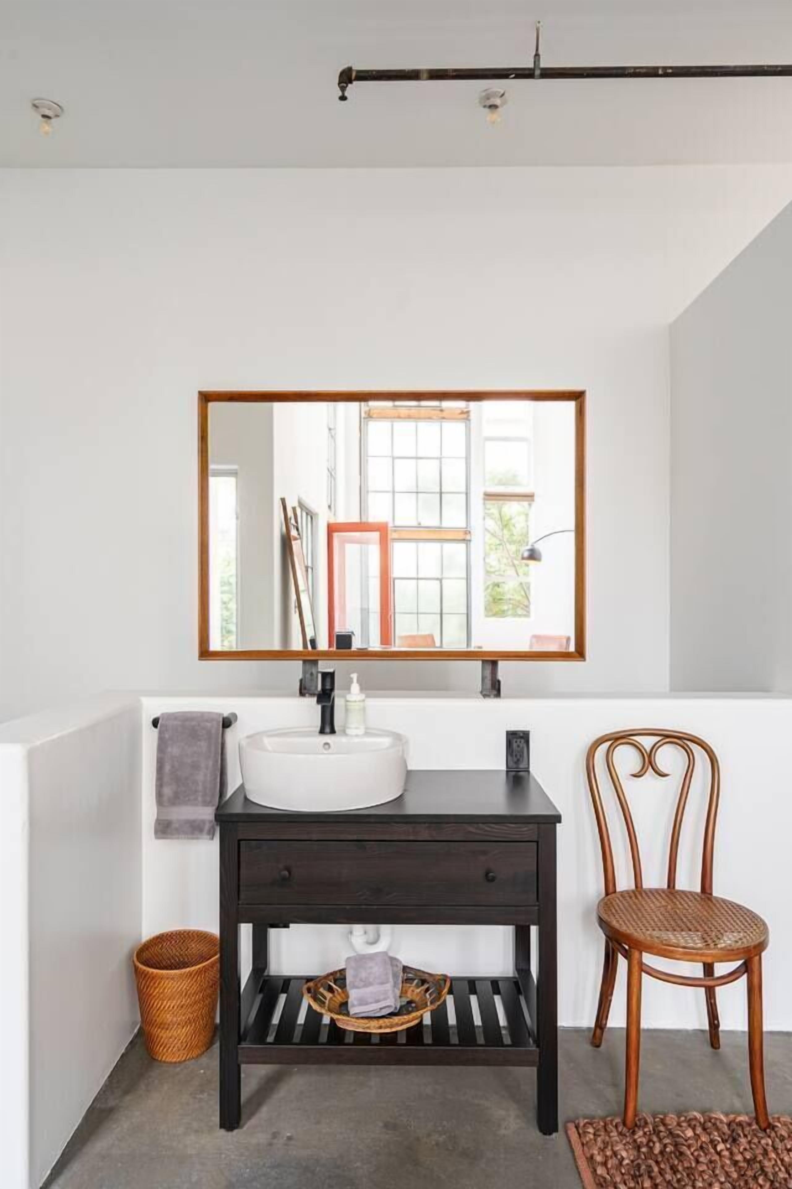 Une salle de bain avec un lavabo blanc, un meuble-lavabo en bois foncé, un miroir et une chaise en osier.