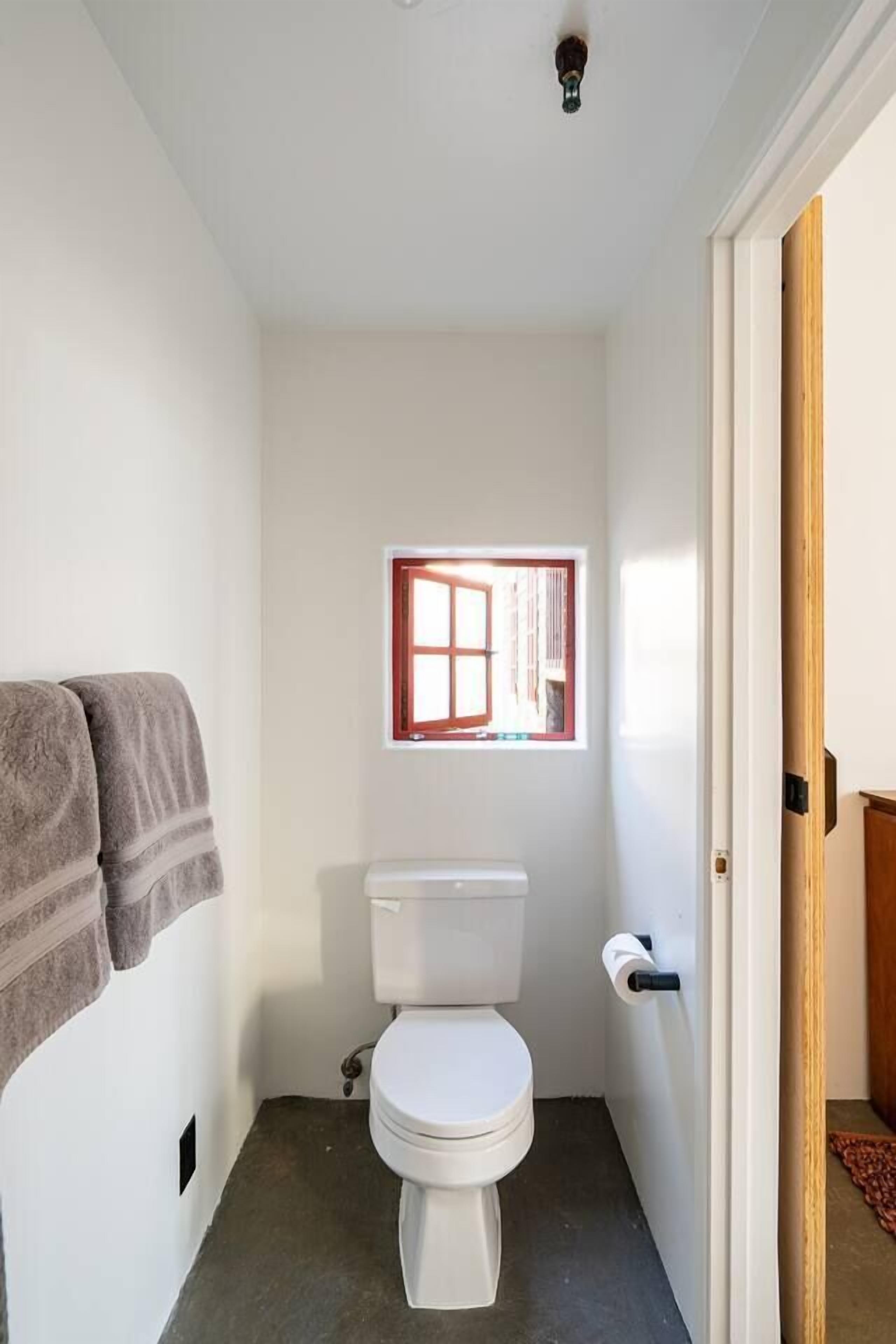Un baño pequeño y funcional con un inodoro blanco, una ventana con marco rojo y una toalla colgada en la pared.