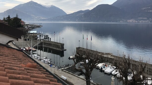 A lakeside view with mountains in the background, a dock with boats, and a building with a sloped roof. - Lake Sunset View - Como Lake (Bellano)