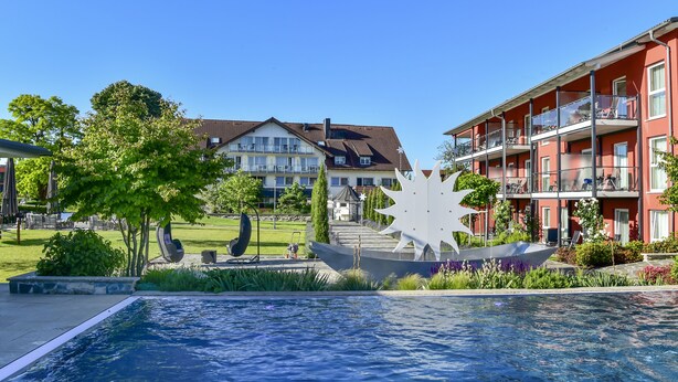 Uma piscina com uma escultura moderna em formato de sol, cercada por vegetação exuberante e um edifício vermelho com varandas.