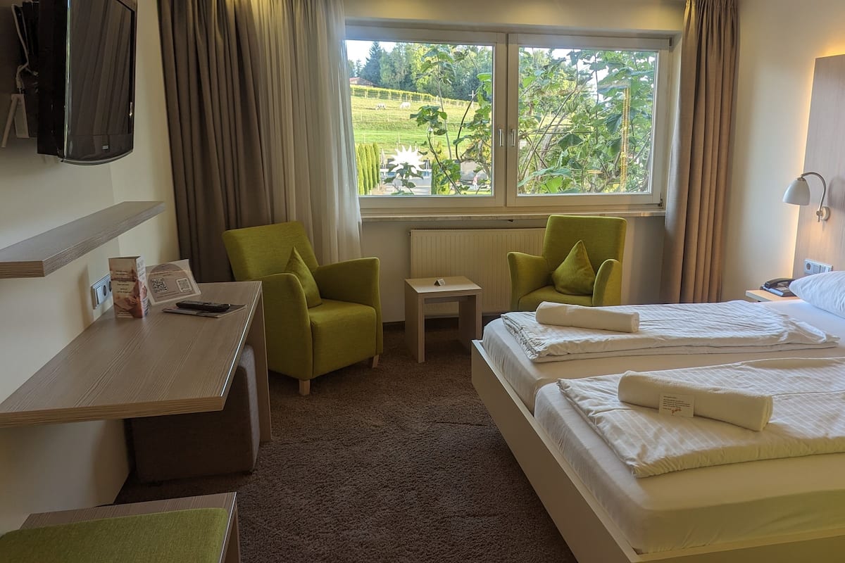 Une chambre d’hôtel avec un lit, deux fauteuils verts, une petite table, un téléviseur et une fenêtre donnant sur un paysage verdoyant.