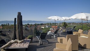 Um terraço com móveis de vime, guarda-sóis brancos e vista para um lago e montanhas.
