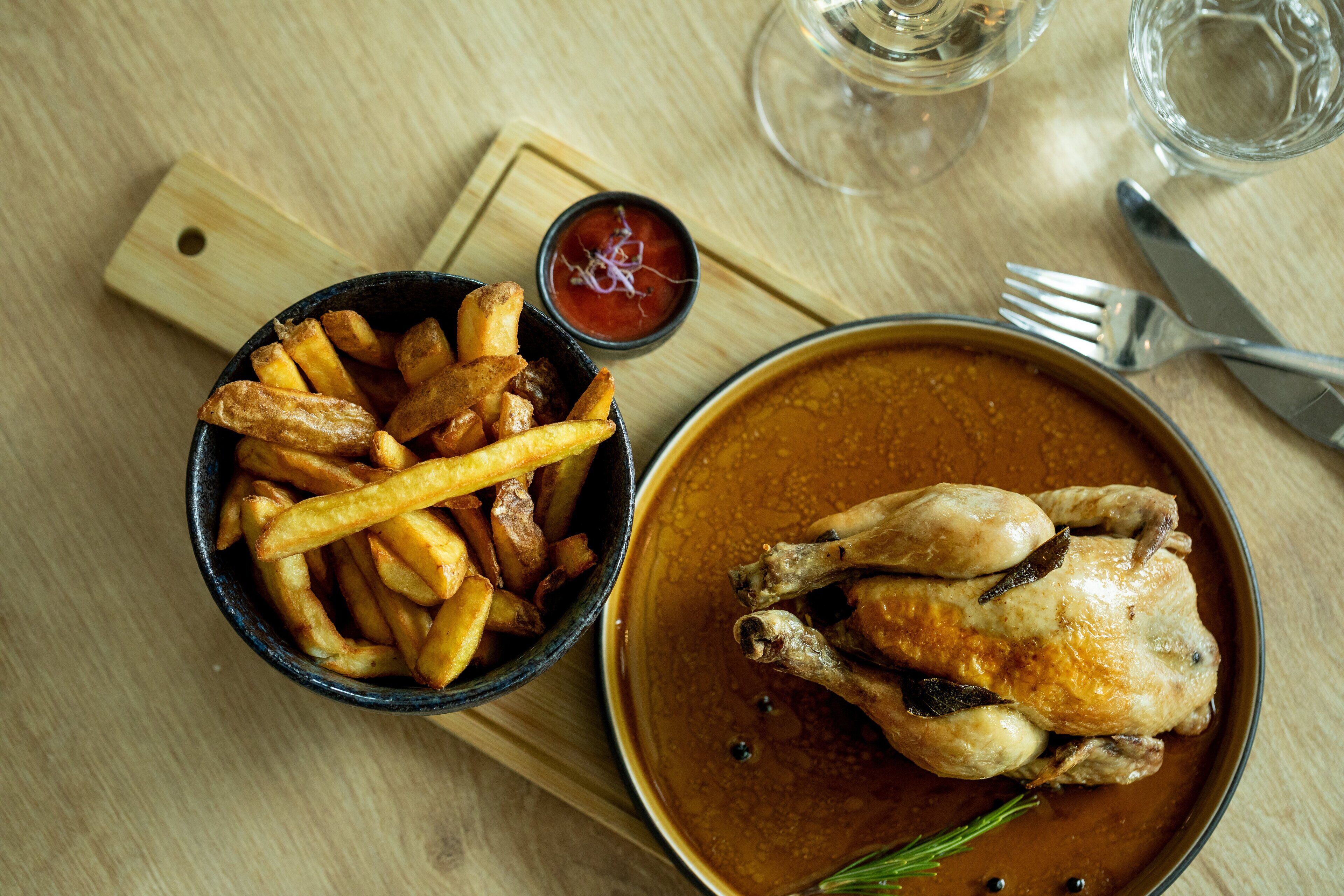 Ein Gericht mit gebratenem Hähnchenschenkel und Pommes, serviert auf einem Holztisch, zusammen mit einem Glas Wasser und einer Gabel.