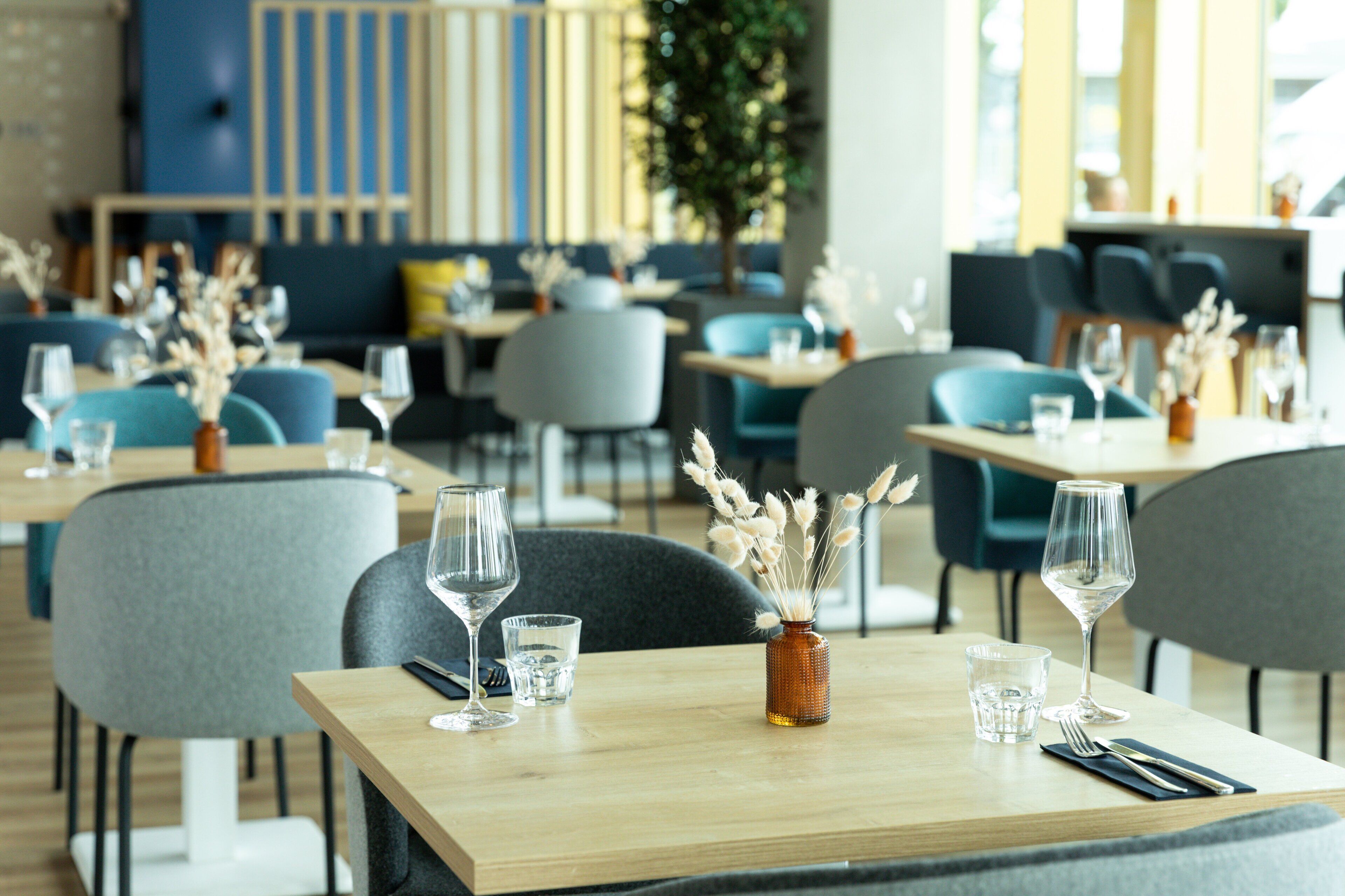 A restaurant with tables set for dining, chairs, and a vase with dried plants.