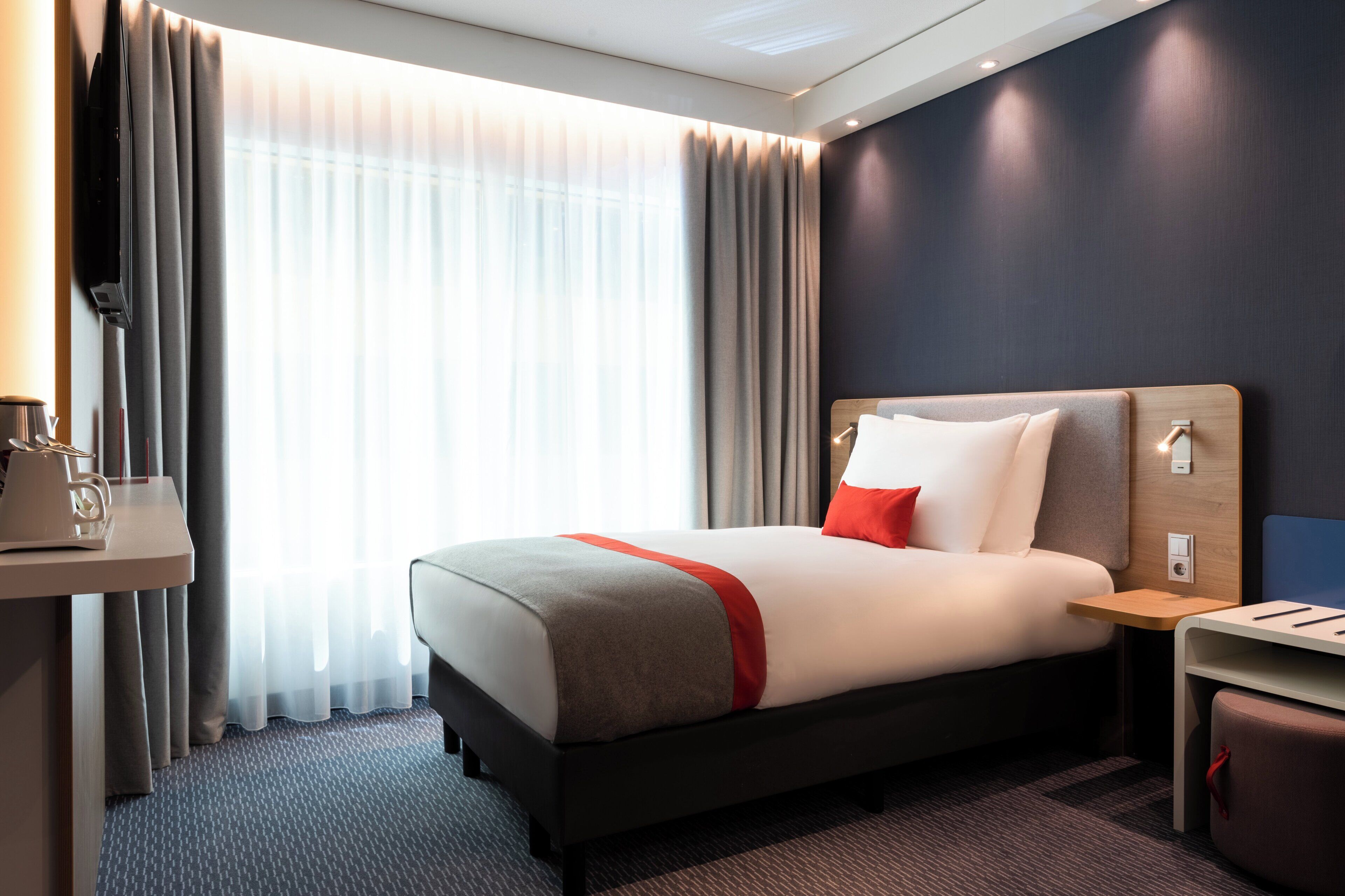 A modern hotel room with a large bed, a bedside table, a desk with a chair, and a large window with curtains.