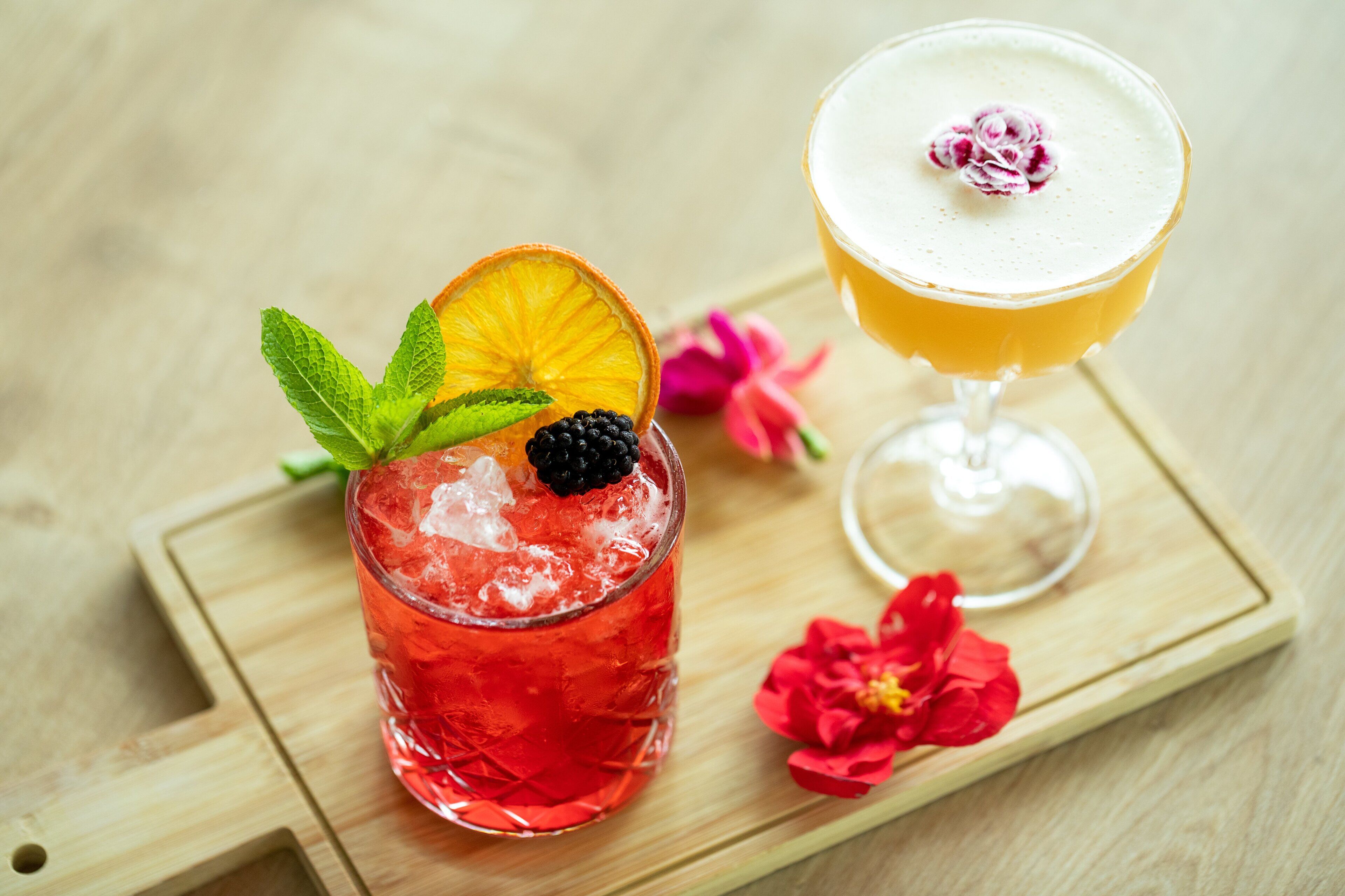 Two cocktails on a wooden tray with a red flower and mint leaves.